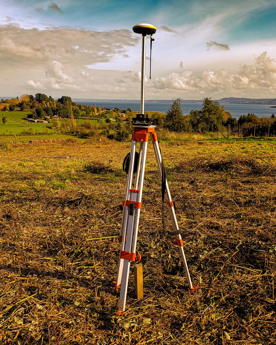 Dos receptores GNSS sobre trípodes en un predio despejado, con vista al lago y cielo de nubes.