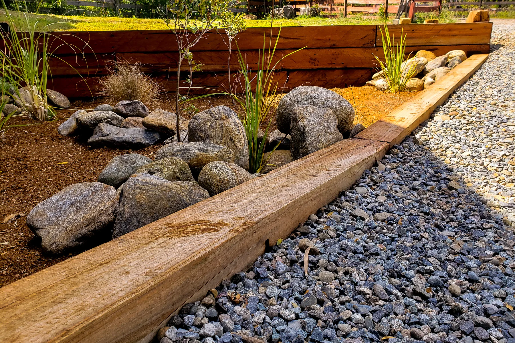 Jardín terminado con piedras de río, gravilla, borde de madera y calle-calle recién plantados bajo arrayanes.