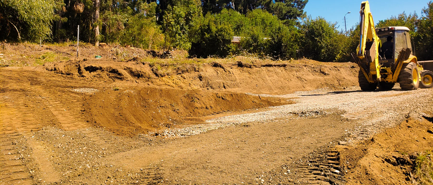 Excavadora moviendo tierra en una ladera sobre el lago, con el bosque al fondo.