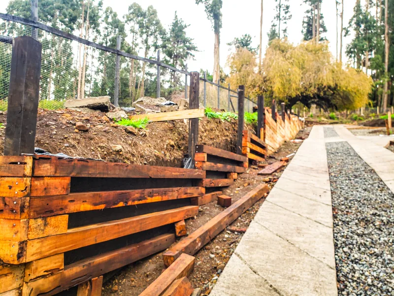 Muro de madera recién construido junto a un sendero de gravilla, entre árboles.