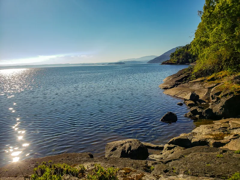 Orilla de piedra del lago con el agua brillando al sol.