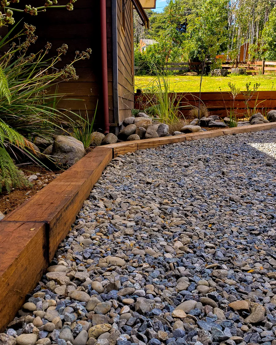 Sendero de gravilla entre macizos con borde de madera, helechos y gramíneas, junto a un muro de madera.